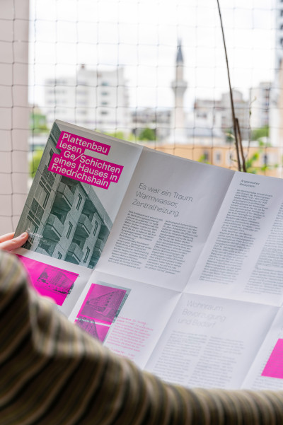 Eine Person hält den Flyer in den Händen. Im unscharfen Hintergrund sind Plattenbauten zu erkennen.