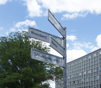 Aluminiummast mit vier Schildern vor einem blauen Himmel mit weißen Wolken: Neuruppin 14 Stunden 2 Minuten, verdeckt vom nächsten Schild: Rheinsbe… , Wustrau 12 Stunden 3 Minuten, Schloss Oranienburg 9 Stunden