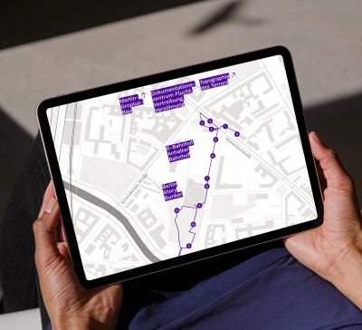 Hände halten ein Tablet. Auf dem Tablet ist eine Seite der Website zu sehen, die die Karte rund um den Anhalter Bahnhof und die verschiedenen Stationen der Audiotour zeigt.