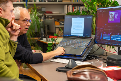 Zwei Männer sitzen an einem Schreibtisch vor einem Laptop und blicken konzentriert auf den Bildschirm, auf dem ein Textdokument geöffnet ist. Der Mann im Vordergrund mit Brille und grauen Haaren tippt auf dem Trackpad, der andere Mann daneben stützt nachdenklich das Kinn auf die Hand. Rechts steht ein zweiter Monitor mit einer Videobearbeitungssoftware, im Hintergrund sind Bücherregale und Zimmerpflanzen zu sehen.